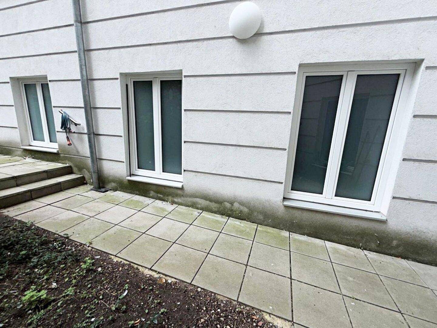 Außenansicht einer Gebäudewand mit drei Fenstern, einem gepflasterten Weg und Stufen zum Eingangsbereich.
