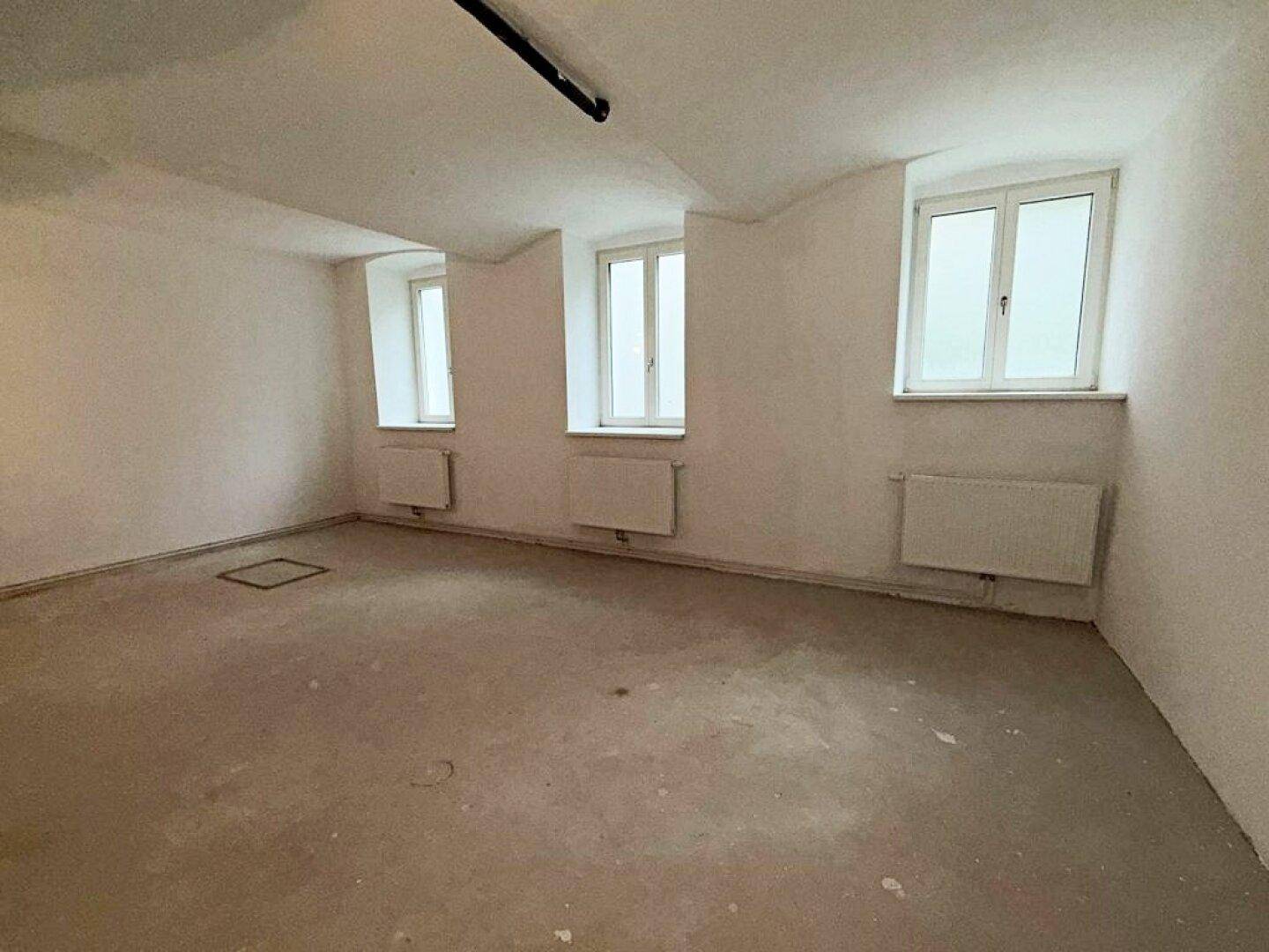 Heller Kellerraum mit Betonboden, weißen Wänden, Heizkörpern und drei Fenstern für natürliche Belichtung.