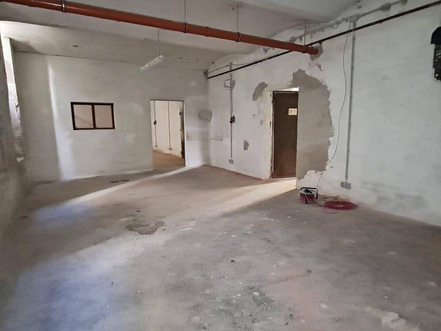 Großer Kellerraum mit Betonboden, sichtbaren Rohrleitungen und mehreren Türen zu angrenzenden Bereichen, unrenoviert.