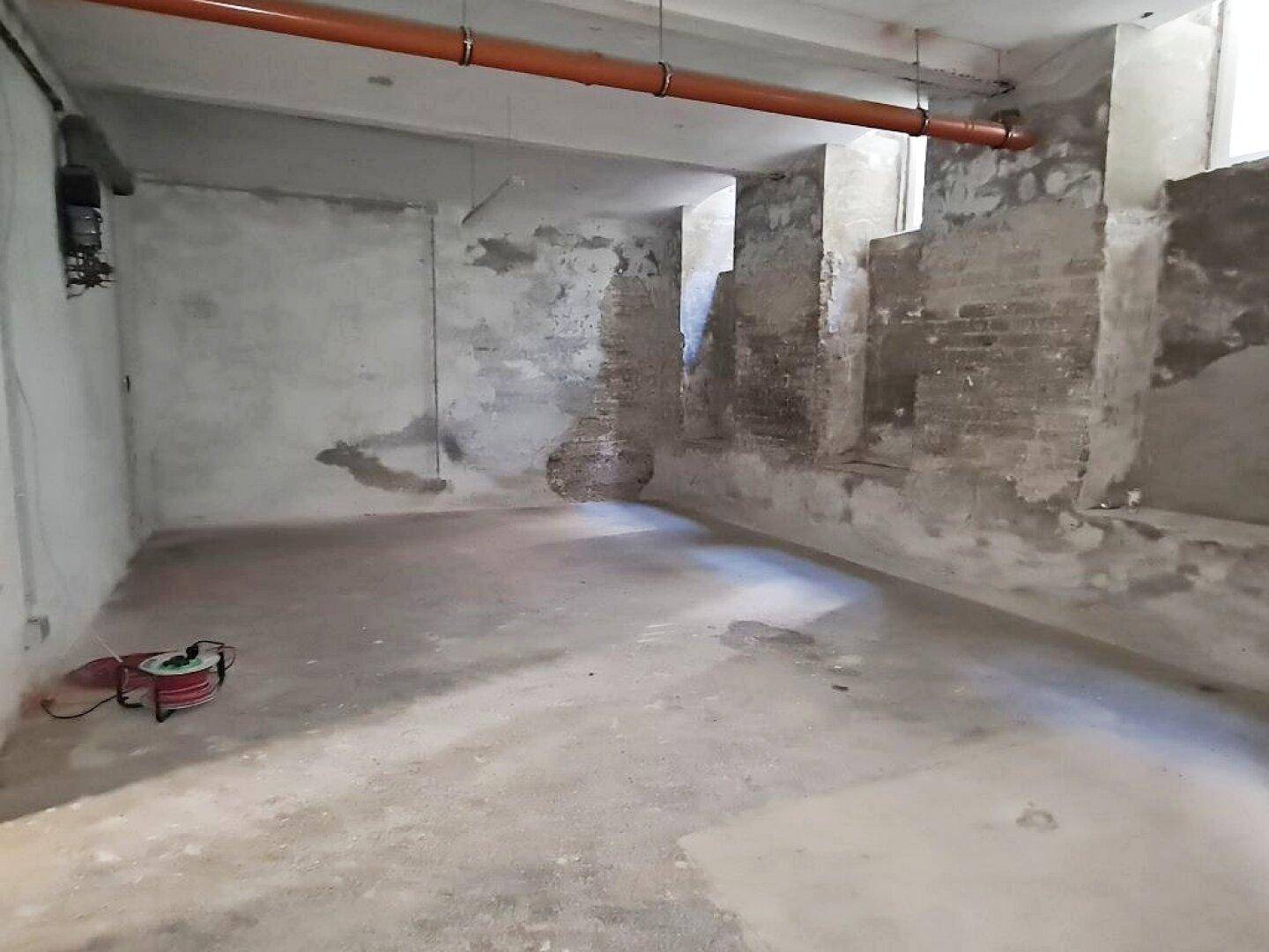 Kellerraum mit unverputzten Wänden, Betonboden und sichtbaren Rohrleitungen an der Decke, in ursprünglichem Zustand.