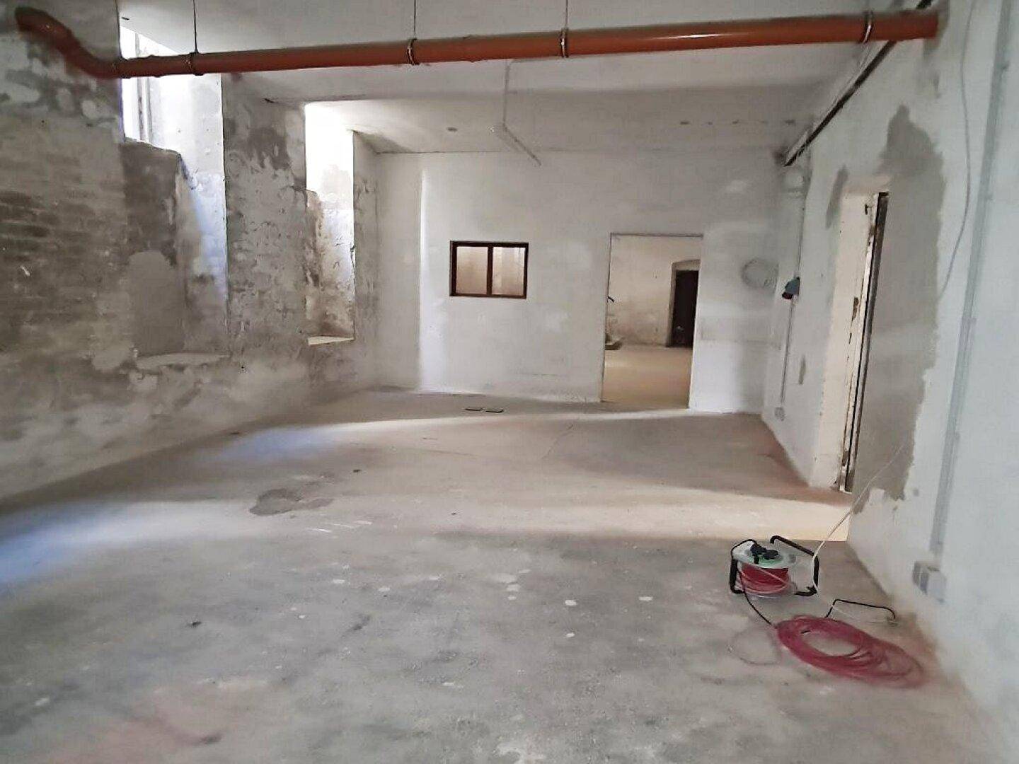 Geräumiger Kellerbereich mit Betonboden, unverputzten Wänden und Zugang zu weiteren Räumen, mit sichtbaren Rohren.