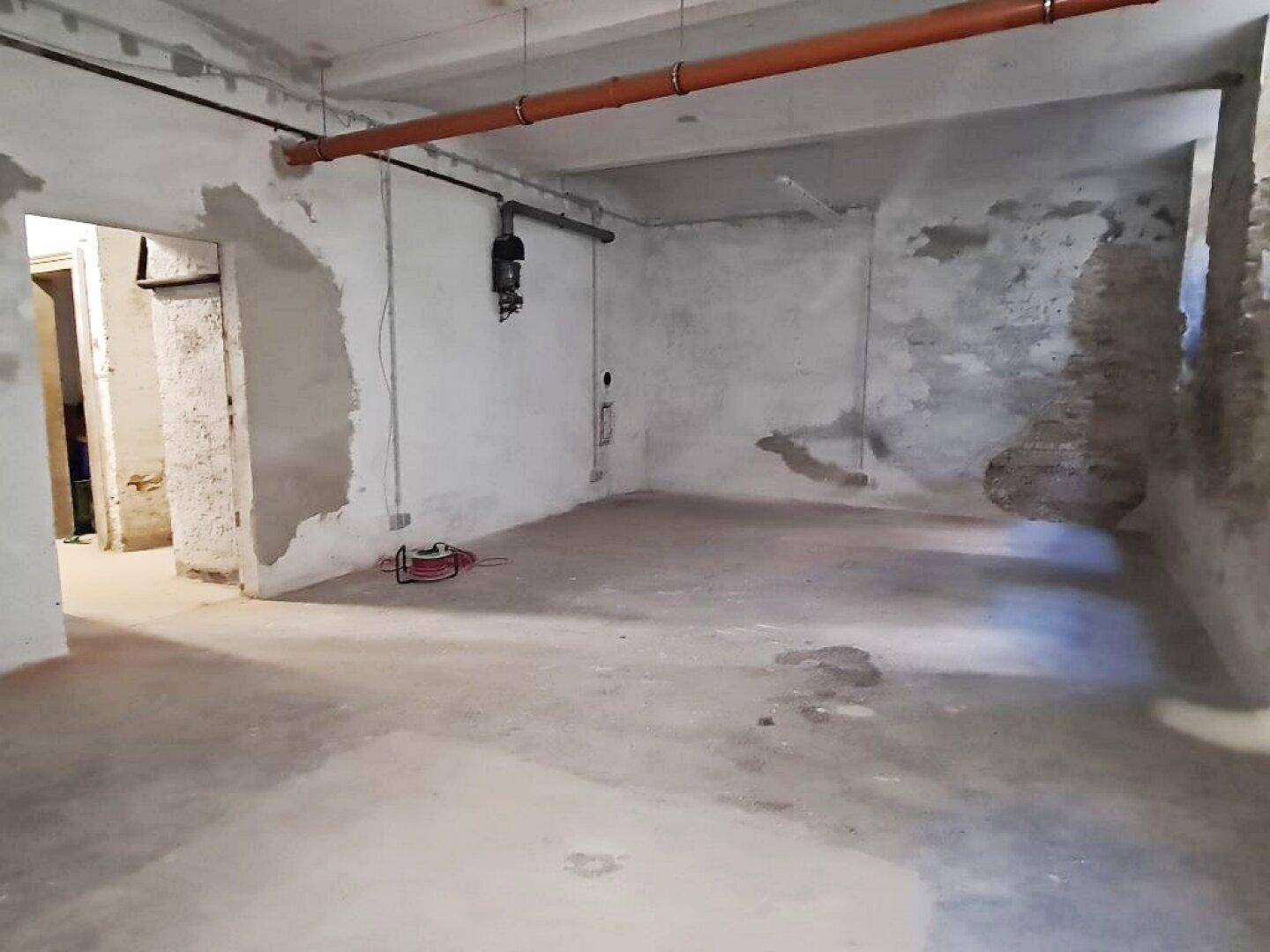 Kellerraum mit Betonboden, unverputzten Wänden und sichtbaren Installationen an der Decke, in sanierungsbedürftigem Zustand.