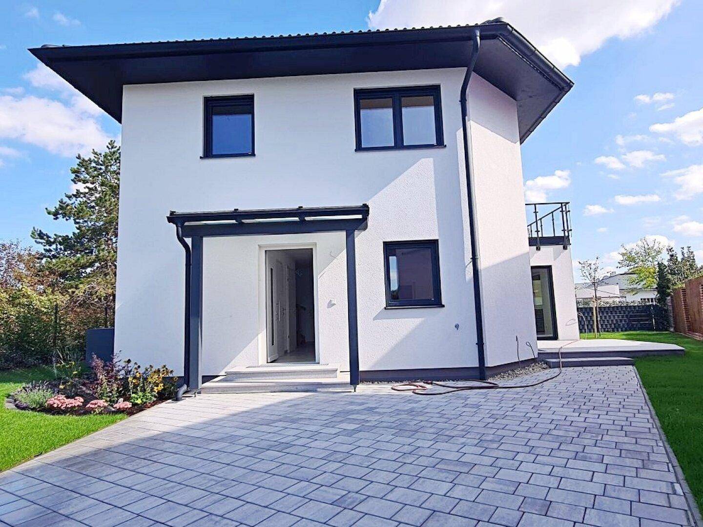 Modernes Einfamilienhaus mit weißer Fassade, dunklem Dach und gepflasterter Einfahrt.
