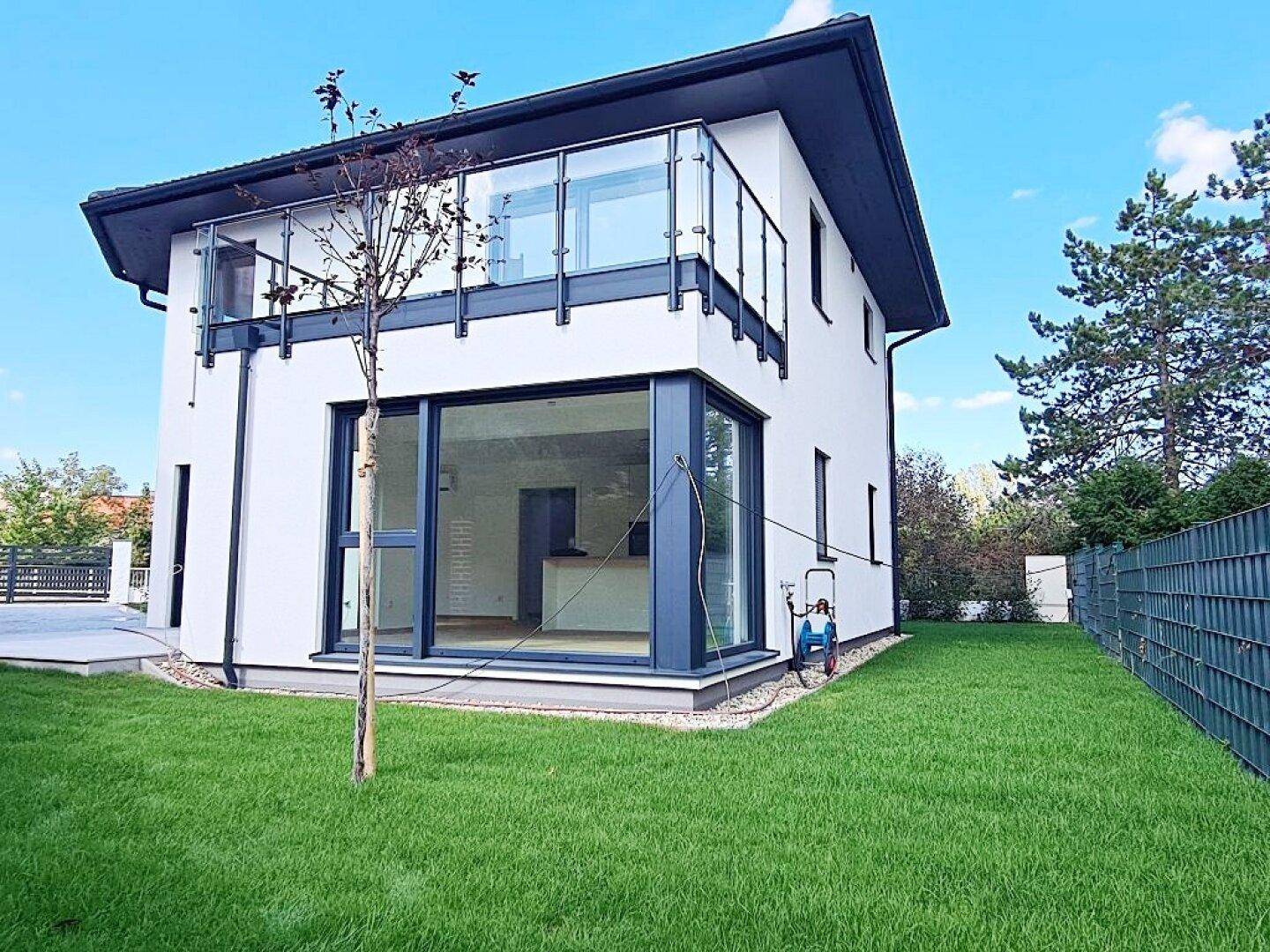 Seitenansicht des modernen Hauses mit großen Fenstern, Balkon und grünem Rasenbereich.