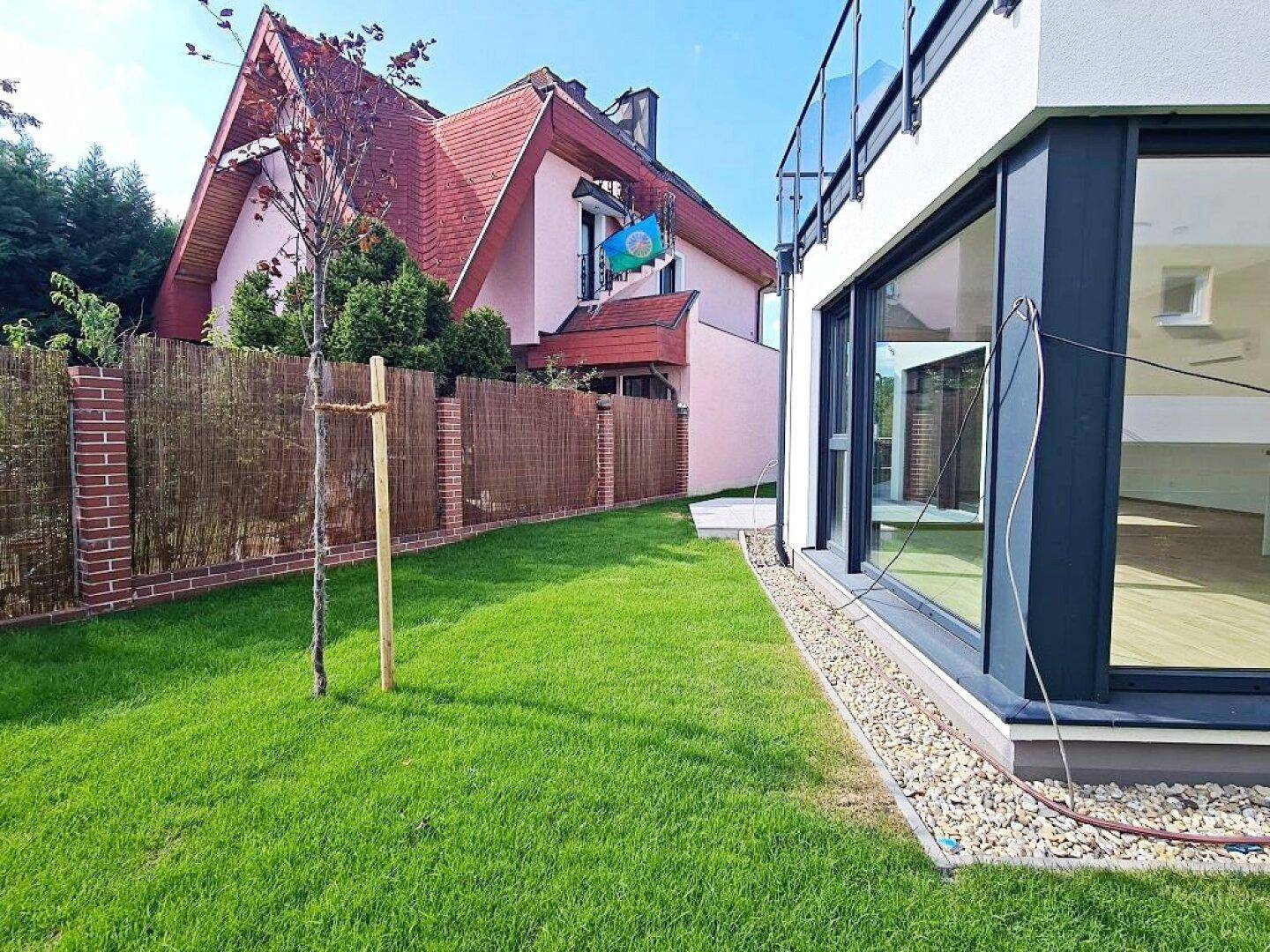 Grüner Gartenbereich neben dem Haus mit großen Fenstern und einem Zaun.