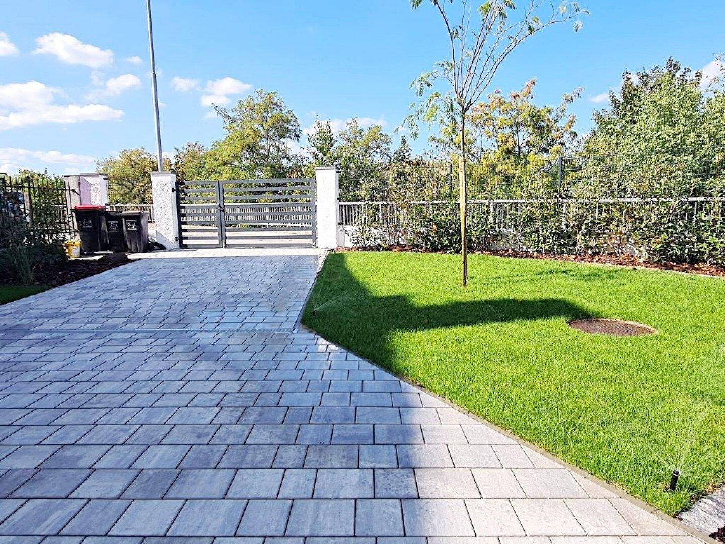 Vorderer Gartenbereich mit gepflasterter Einfahrt, Rasenfläche und einem modernen Tor.