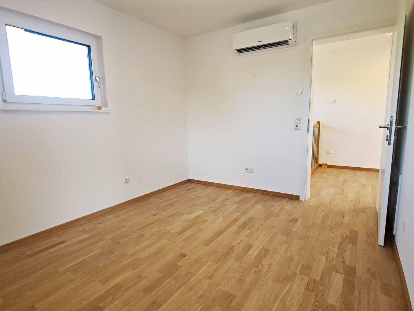 Geräumiges Zimmer mit Klimaanlage und Parkettboden, verbunden mit einem hellen Flur.