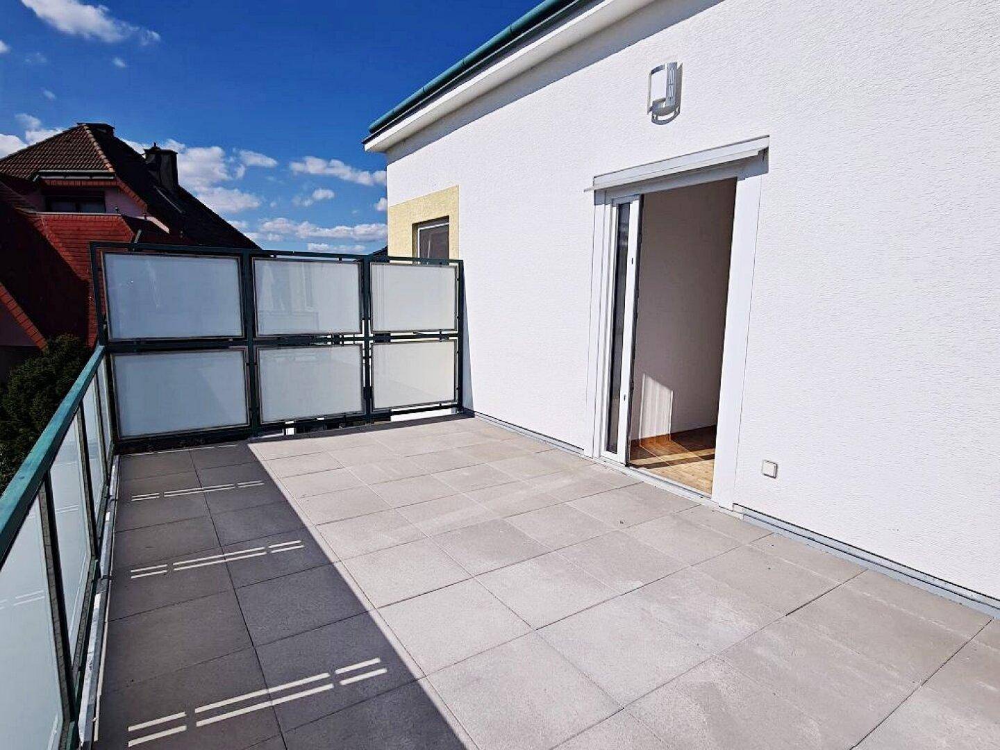 Helle Terrasse mit Fliesenboden und moderner Glasbrüstung, mit Zugang zu einem Innenraum.