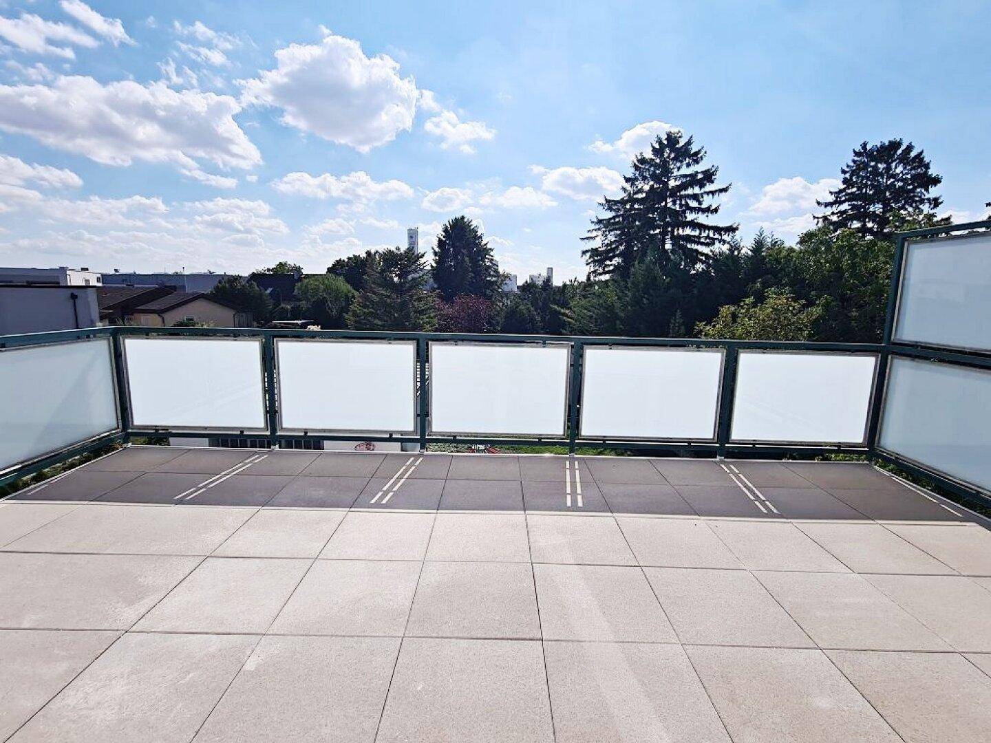 Helle Terrasse mit Fliesenboden und Glasbrüstung, bietet einen weiten Blick auf die Baumlandschaft.