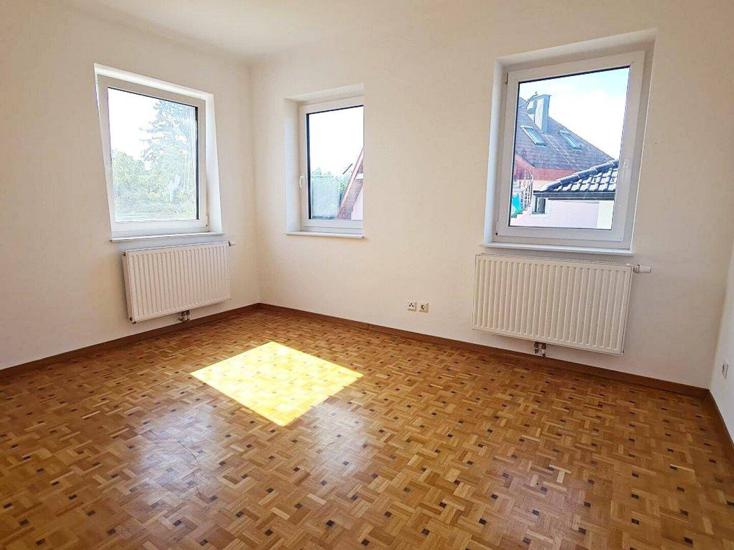 Leeres Zimmer mit klassischem Parkettboden und mehreren Fenstern, die viel Licht hereinlassen.