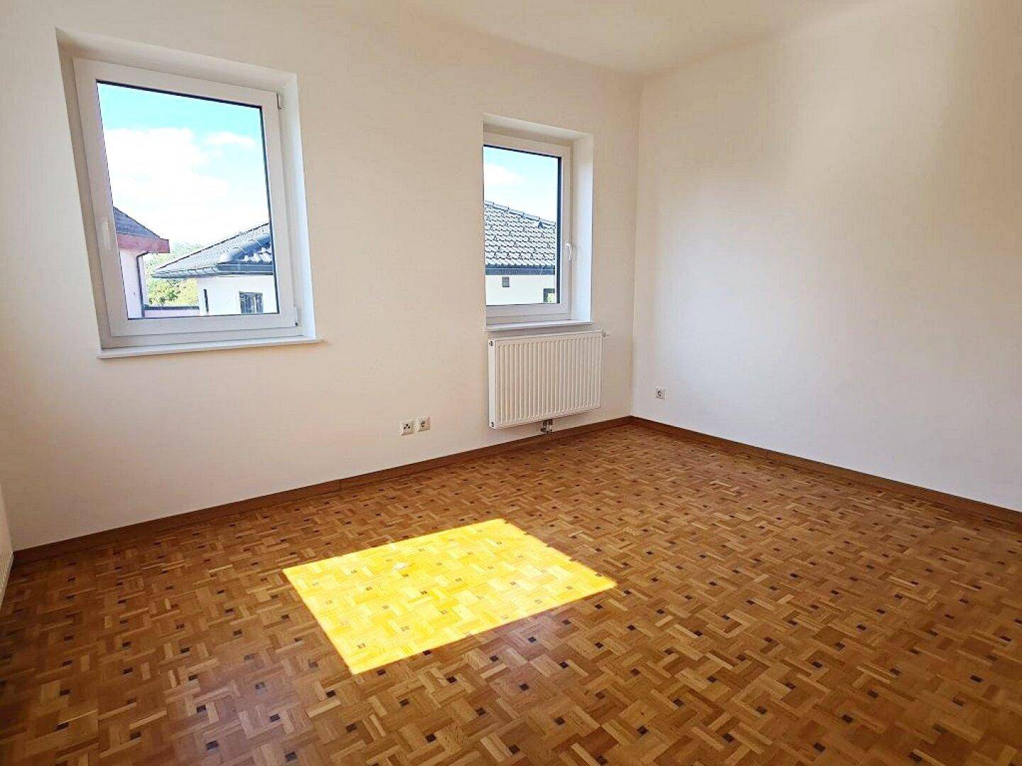 Helles, leeres Zimmer mit Parkettboden und zwei Fenstern, die den Raum sonnig erscheinen lassen.