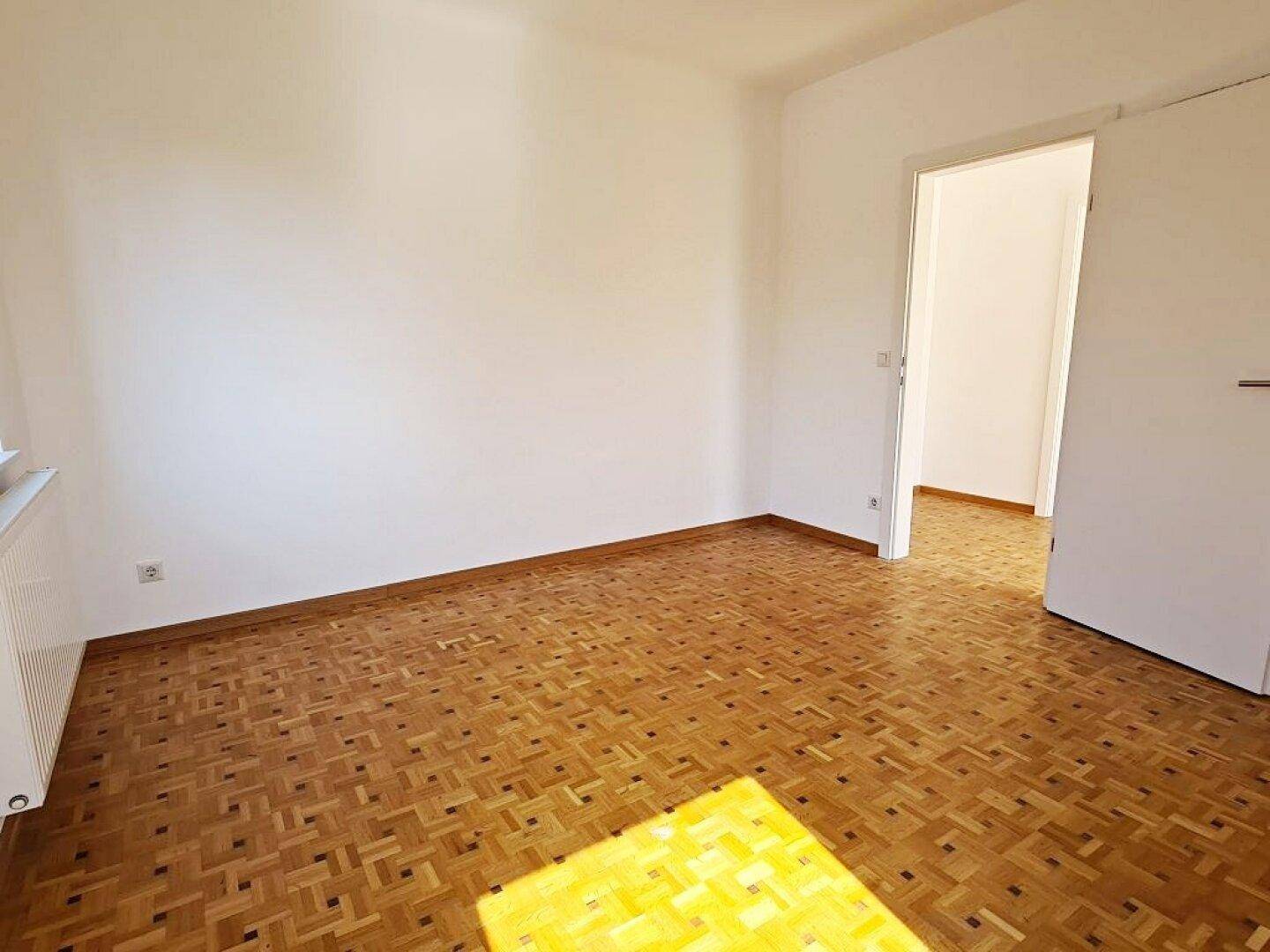 Leeres Zimmer mit Parkettboden und offenem Durchgang zu einem weiteren hellen Raum.