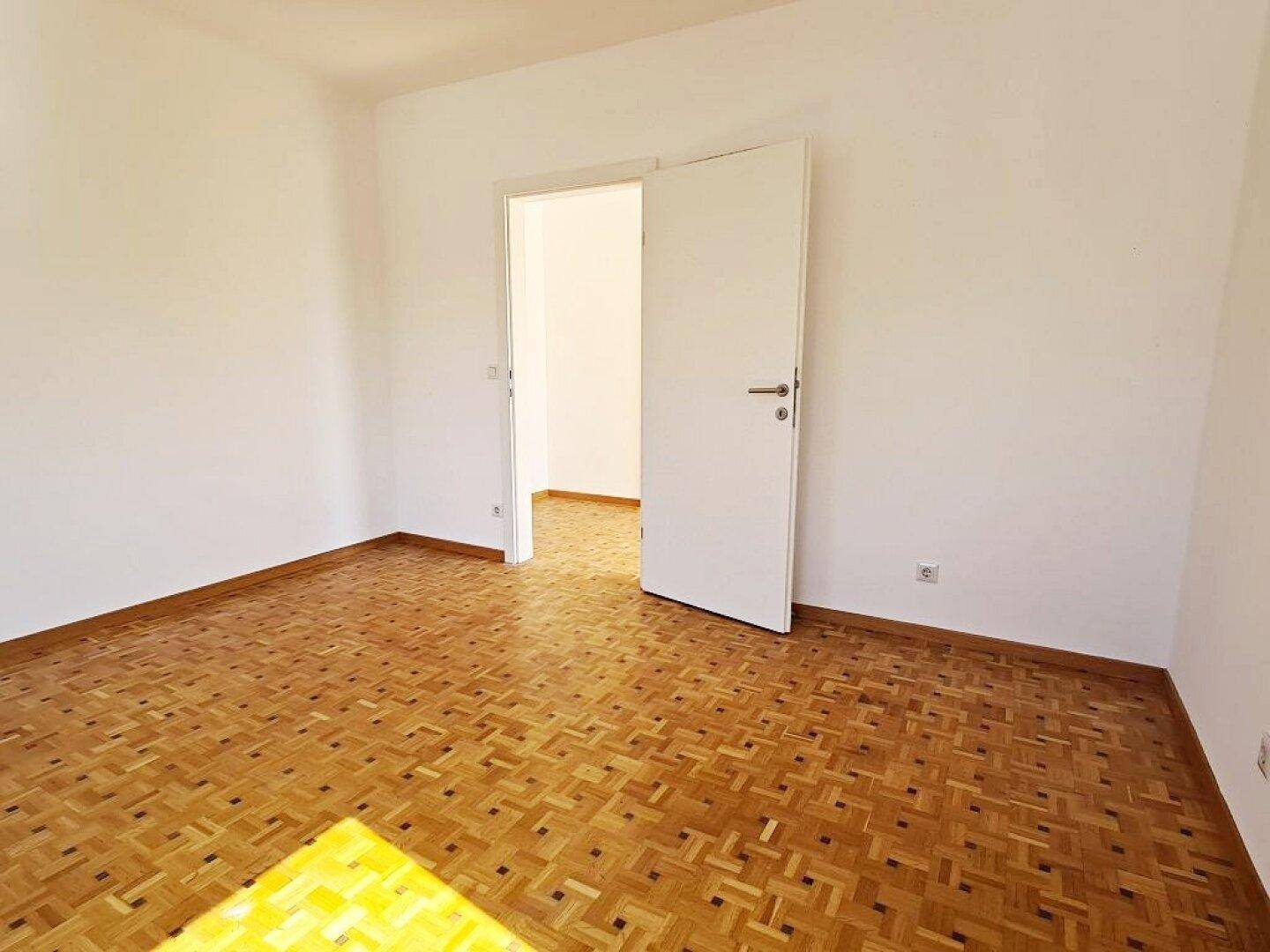 Geräumiges, leeres Zimmer mit Parkettboden und einer offenen Tür zu einem angrenzenden Bereich.