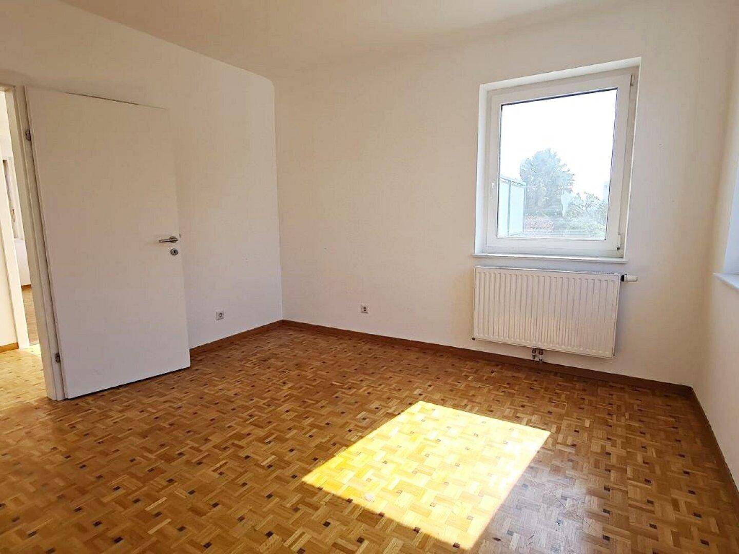 Helles, leeres Zimmer mit Parkettboden und einem Fenster, das Tageslicht hereinlässt.