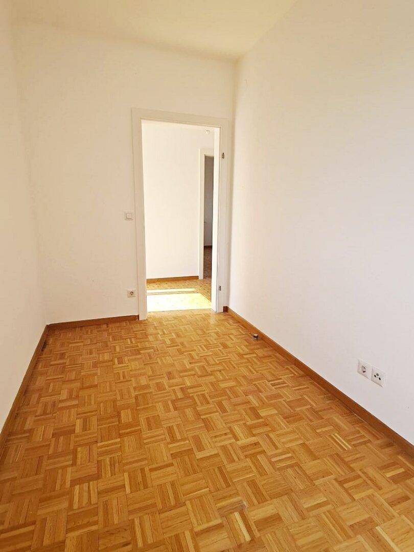 Leeres Zimmer mit Parkettboden und offenem Durchgang zu einem weiteren hellen Raum.