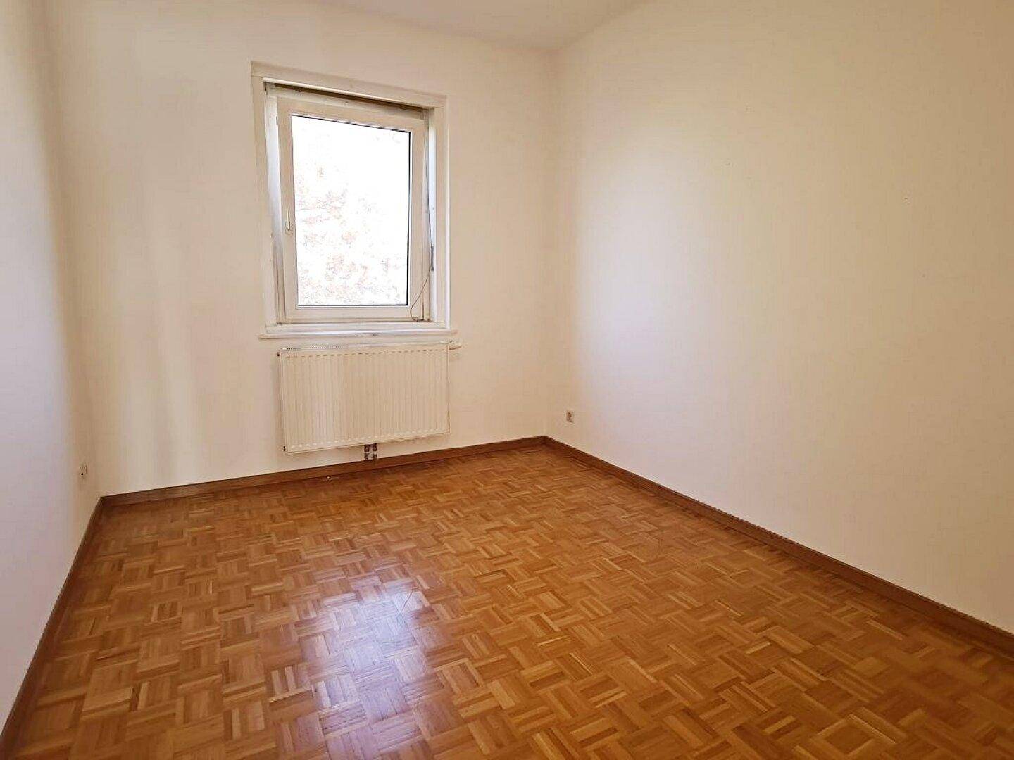 Helles, leeres Zimmer mit Parkettboden und einem Fenster, das den Raum gut belichtet.