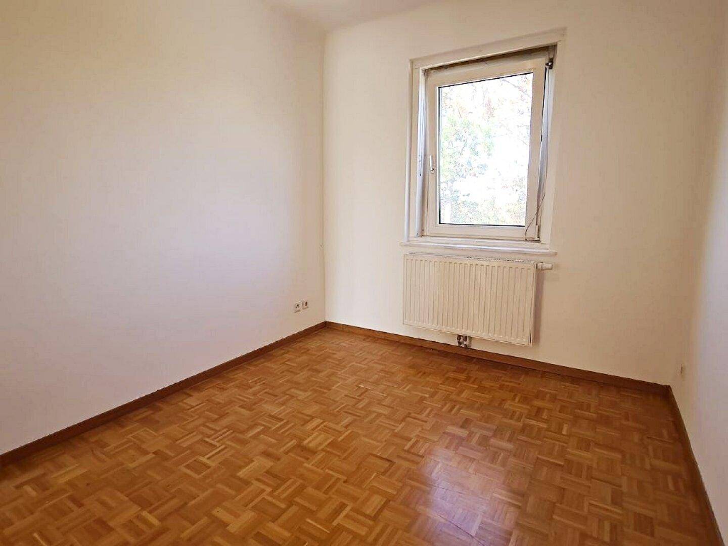Leeres Zimmer mit Parkettboden und einem Fenster, das für ausreichend Helligkeit sorgt.