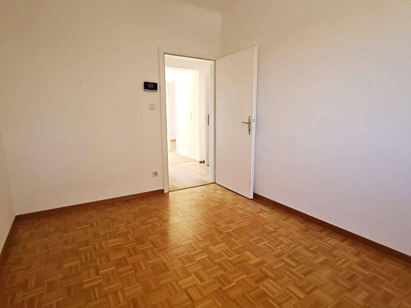 Helles, leeres Zimmer mit Parkettboden und offenem Durchgang zu einem weiteren Bereich.