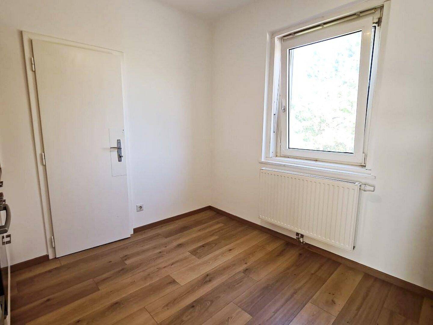 Helles, leeres Zimmer mit Laminatboden und einem Fenster, das viel Licht hereinlässt.