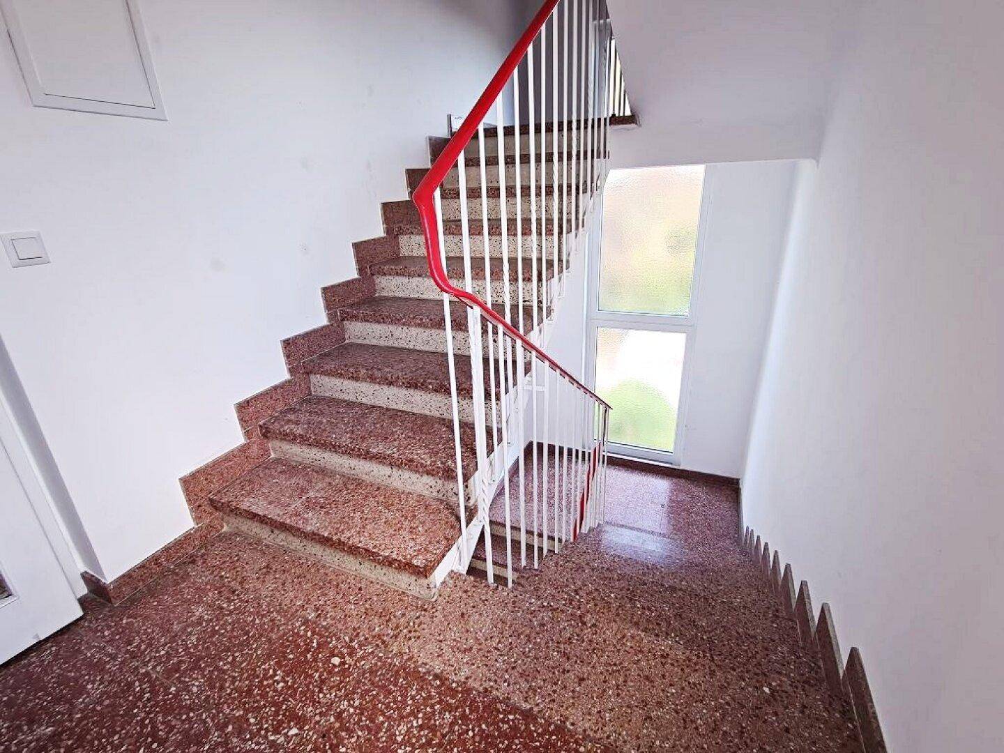 Helles Treppenhaus mit Terrazzostufen und rotem Geländer, das zu den oberen Etagen führt.