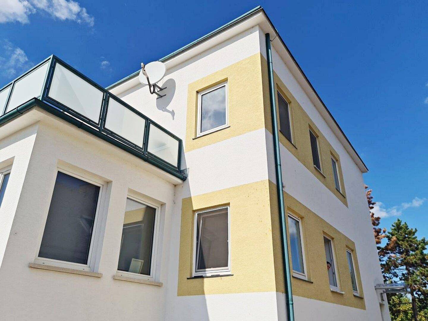 Außenansicht eines Gebäudes mit gelb-weißer Fassade und einem Balkon mit Glasgeländer.