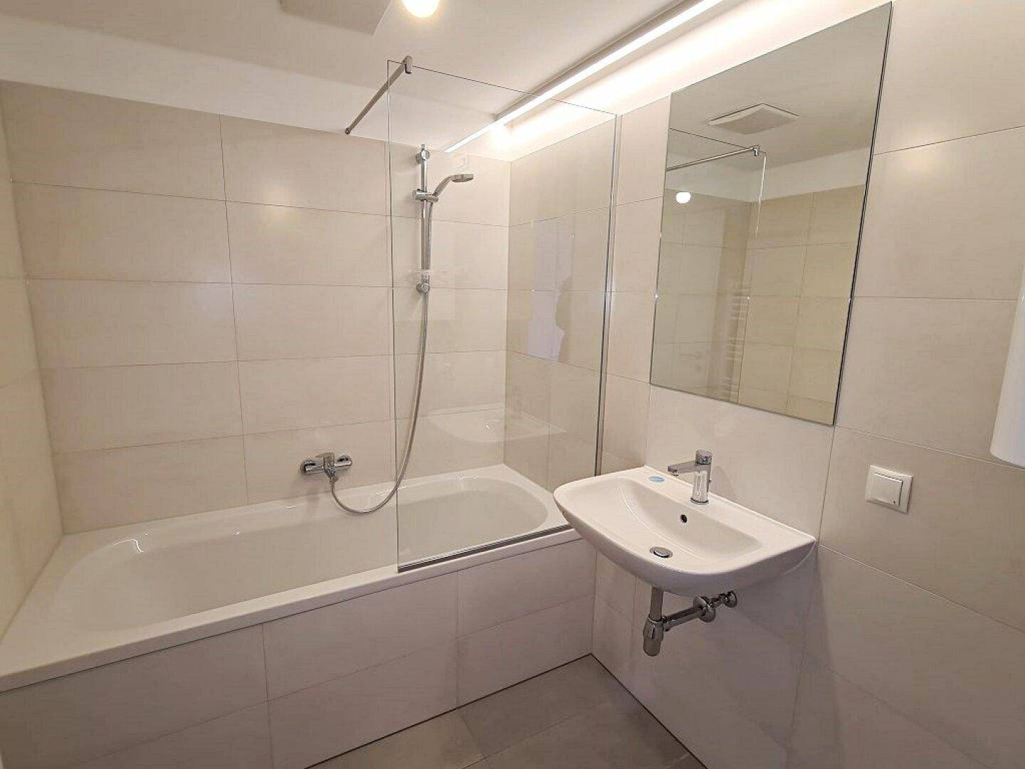 Modernes Badezimmer mit hellgrauen Fliesen, Badewanne mit Duschwand und Waschbecken.