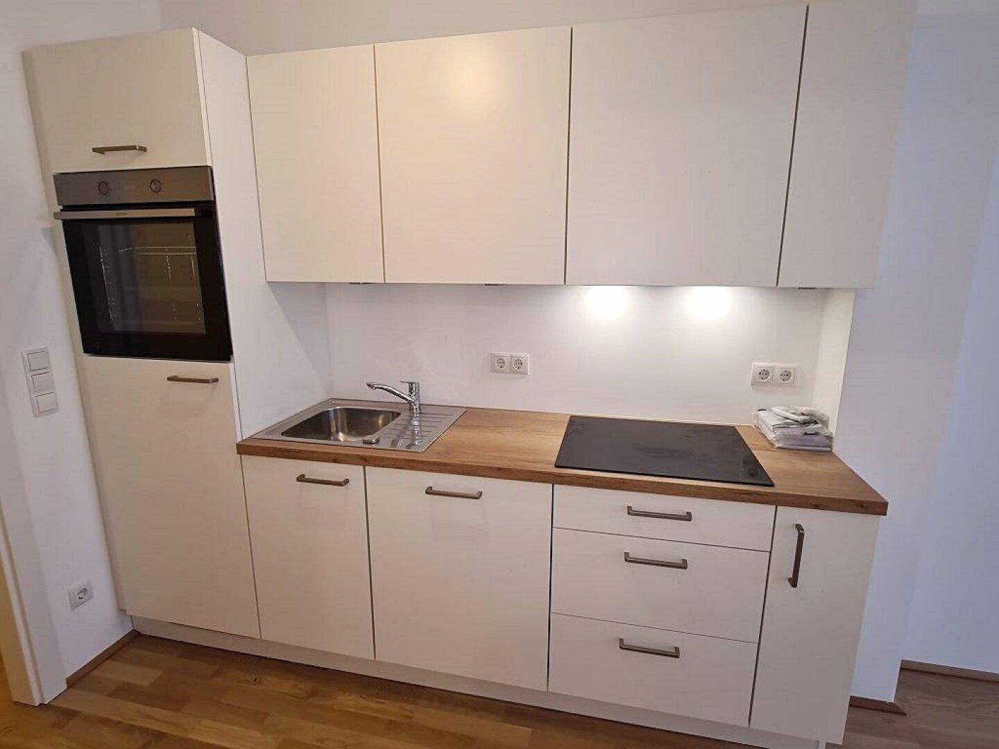 Kompakte, moderne Kochnische mit weißen Fronten, Holz-Arbeitsplatte und integriertem Backofen.
