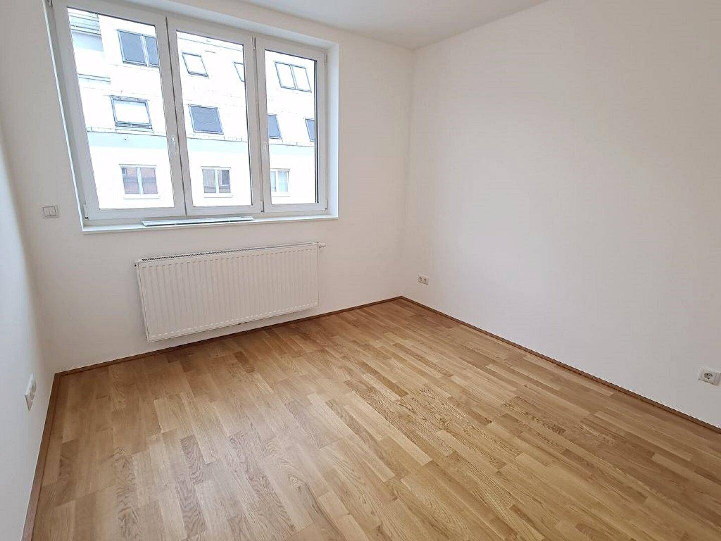 Ruhiges Schlafzimmer mit hellem Parkettboden, weißen Wänden und großem Fenster für viel Tageslicht.