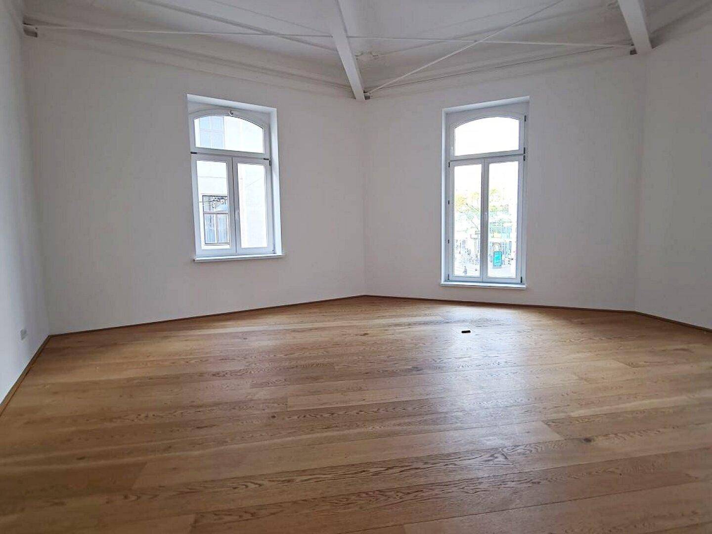 Heller Wohnraum mit elegantem Holzboden, hohen Decken und zwei großen, bogenförmigen Fenstern.