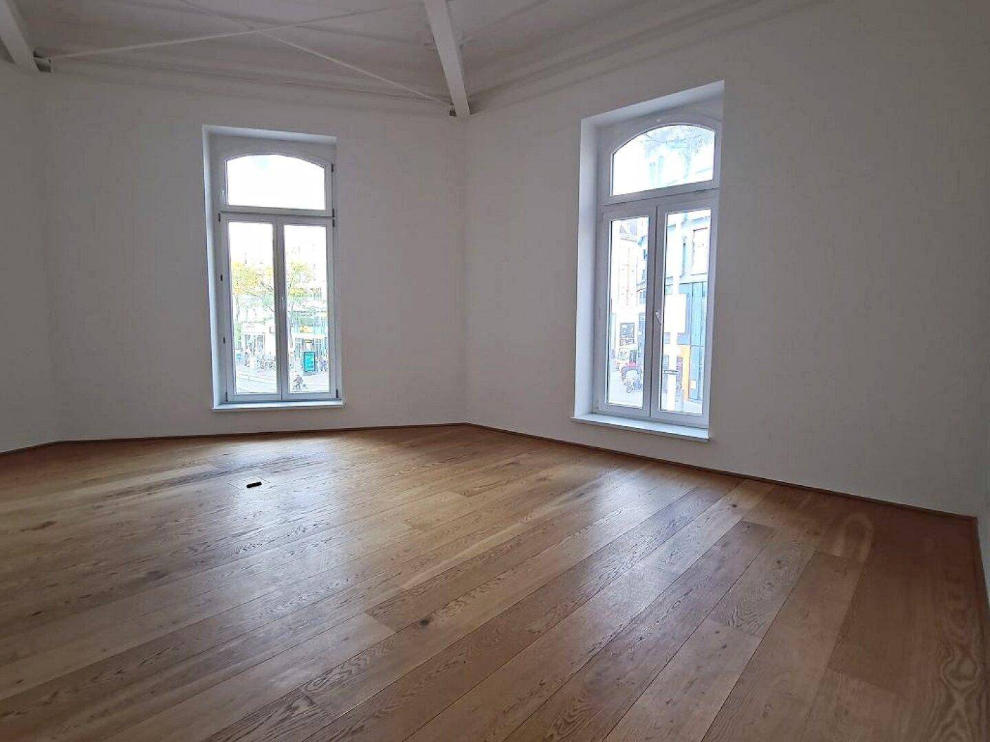 Geräumiger Raum mit hellem Holzboden, hohen Decken und zwei großen Fenstern, die viel Licht hereinlassen.