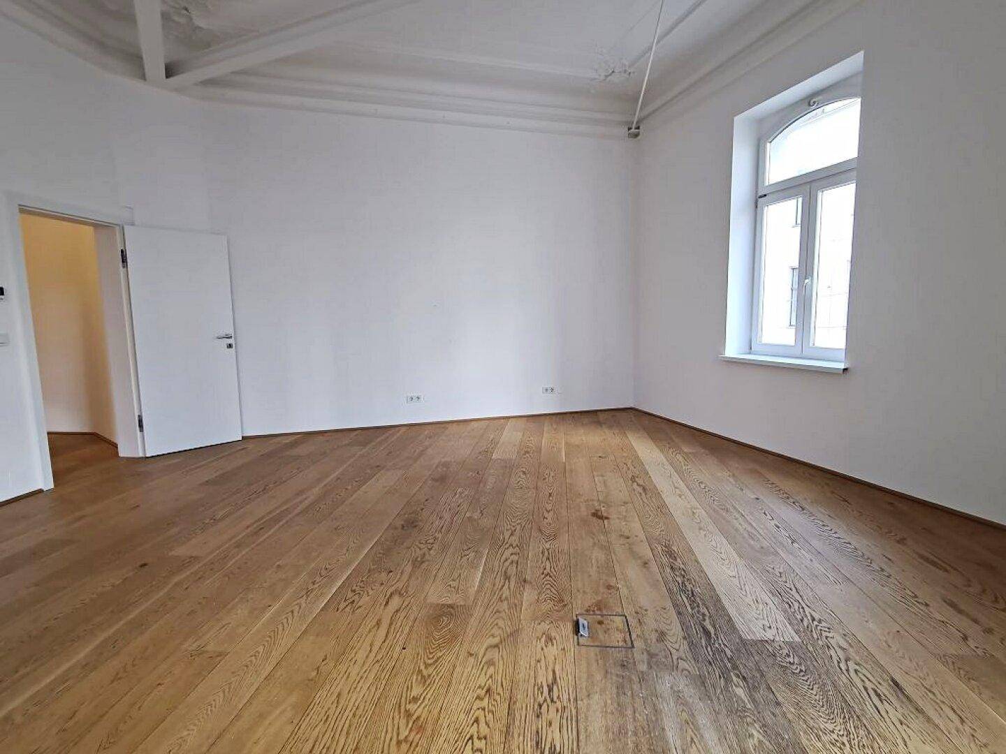 Geräumiger Raum mit elegantem Holzboden, weißen Wänden und einem großen Bogenfenster, das den Raum erhellt.