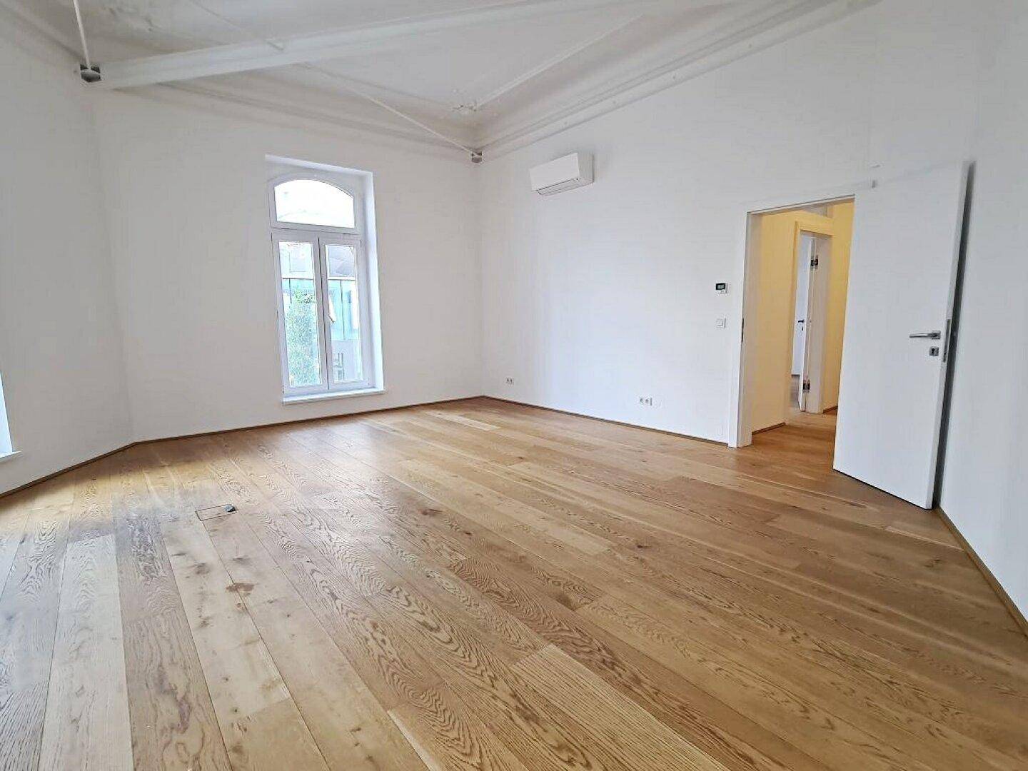 Heller Raum mit Holzboden, weiß gestrichenen Wänden, einem Bogenfenster und Klimaanlage an der Wand.