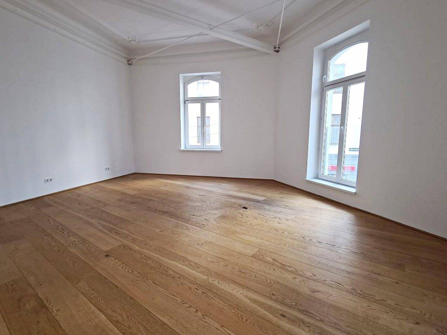 Heller Raum mit Holzboden, hohen Decken und zwei großen Bogenfenstern, die den Raum erhellen.