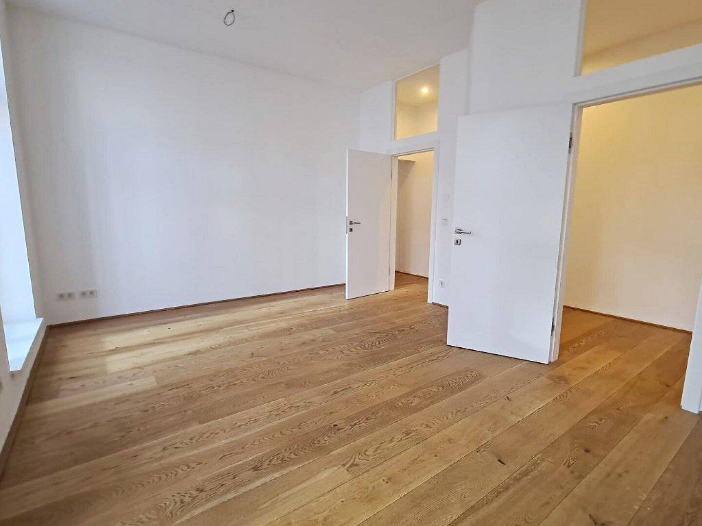 Heller Raum mit Holzboden und zwei offenen Türen, die zu weiteren Zimmern führen.