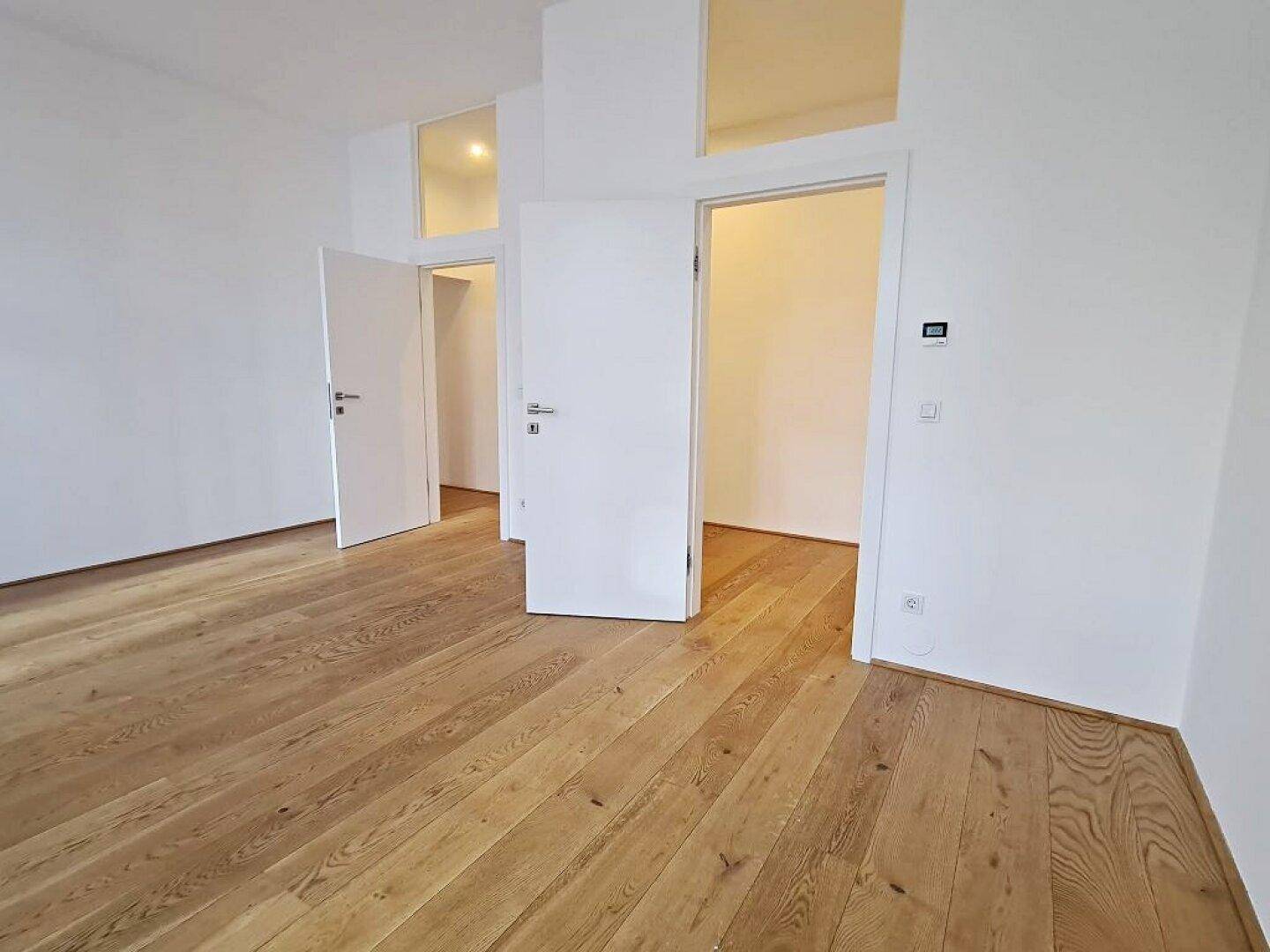 Geräumiger Raum mit Holzboden und zwei offenen Türen, die den Zugang zu angrenzenden Räumen ermöglichen.
