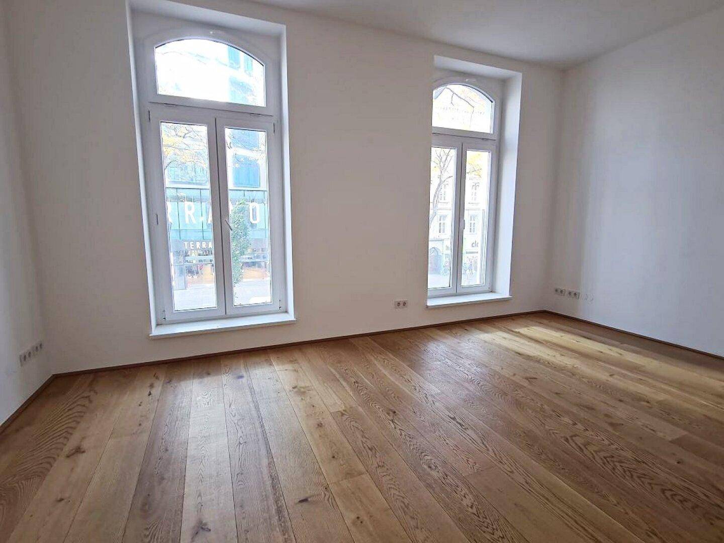 Geräumiger Raum mit Holzboden, weißen Wänden und zwei großen Bogenfenstern mit Blick auf die Straße.