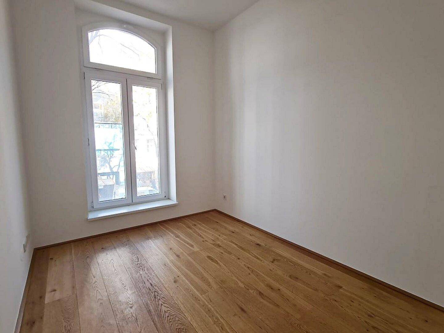 Kleinerer Raum mit Holzboden, weißen Wänden und einem hohen Bogenfenster, das viel Licht spendet.