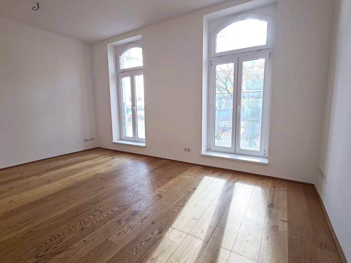 Heller Raum mit Holzboden, weißen Wänden und zwei großen Bogenfenstern, die Sonnenlicht hereinlassen.