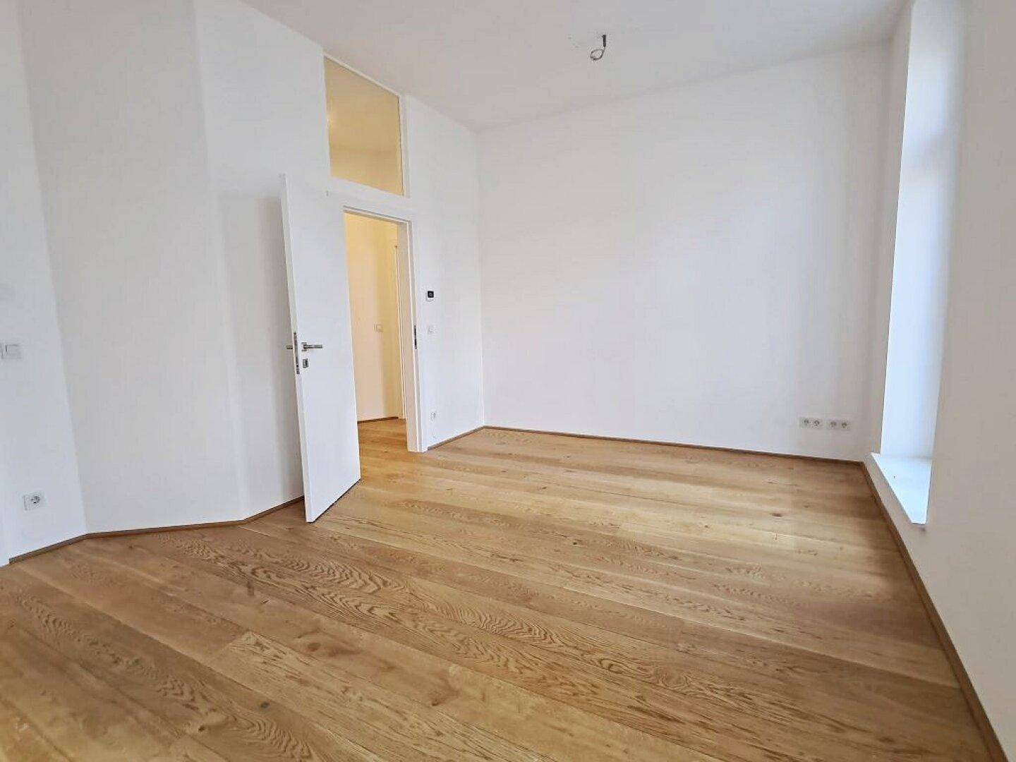 Heller Raum mit Holzboden, weißen Wänden, einem großen Fenster und einer offenen Tür.