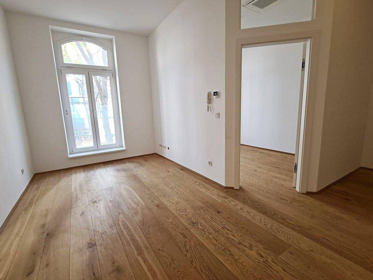 Geräumiger Raum mit Holzboden, weißen Wänden, einem großen Fenster und einer offenen Tür.