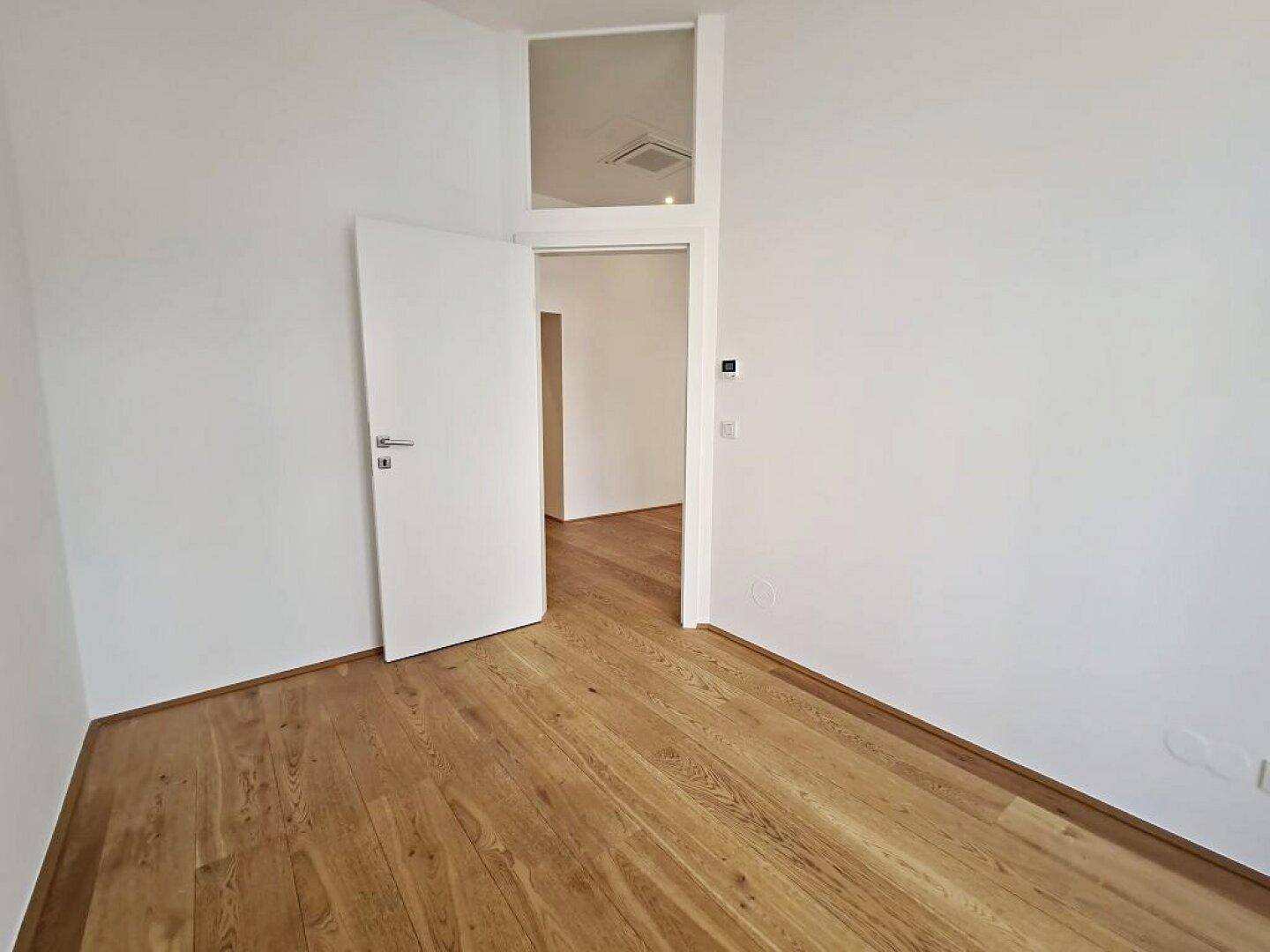 Heller Raum mit Holzboden, weißen Wänden und einer offenen Tür, die zu einem weiteren Zimmer führt.