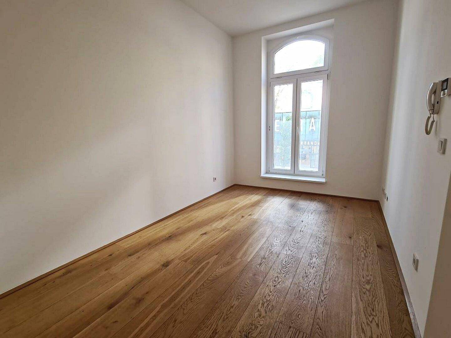 Heller Raum mit Holzboden, weißen Wänden, einem Bogenfenster und einer Gegensprechanlage an der Wand.
