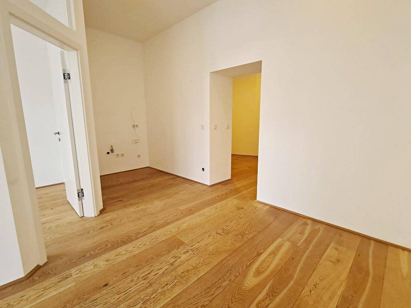 Heller Raum mit Holzboden, weißen Wänden und einer offenen Tür, die zu einem weiteren Zimmer führt.