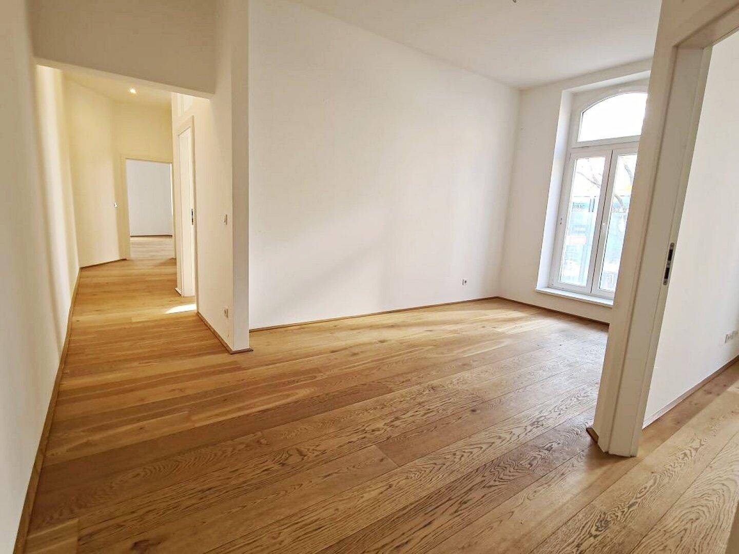 Geräumiger Raum mit Holzboden, weißen Wänden, einem großen Fenster und einer offenen Tür.