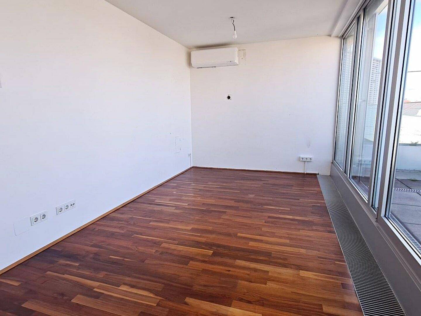 Heller Raum mit Holzboden, großen Fenstern und Klimaanlage, ideal als Wohnzimmer nutzbar.
