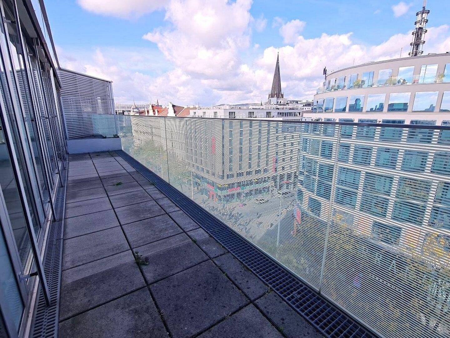 Geräumige Terrasse mit Glasgeländer und weitem Blick über die städtische Umgebung und Gebäude.