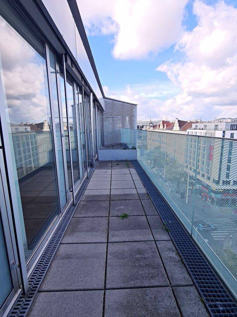 Breite Terrasse mit Glasgeländer und Zugang zu den Innenräumen, mit Stadtblick und Himmel.