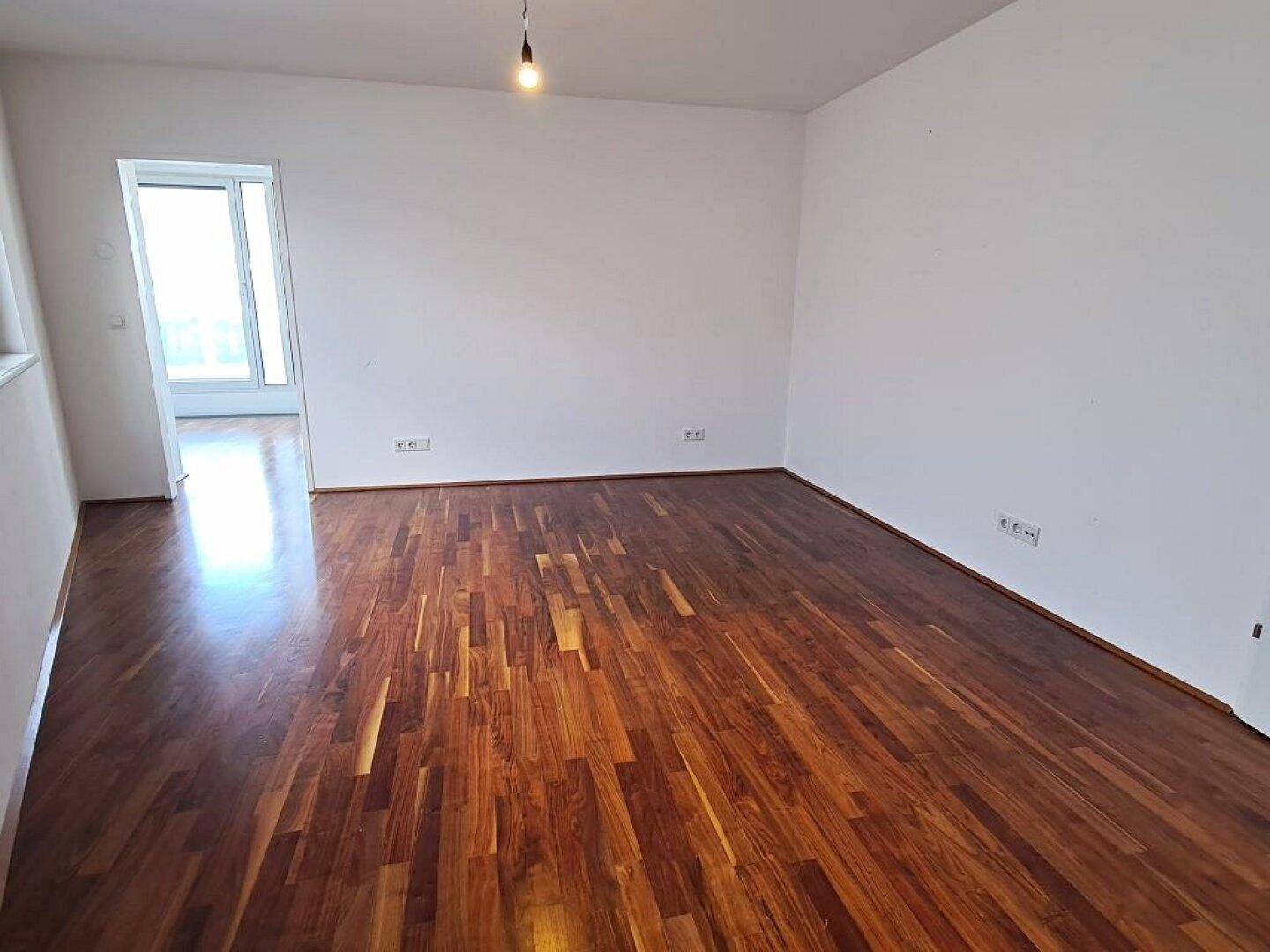 Helles Zimmer mit dunklem Parkettboden und Zugang zu einem weiteren Raum.