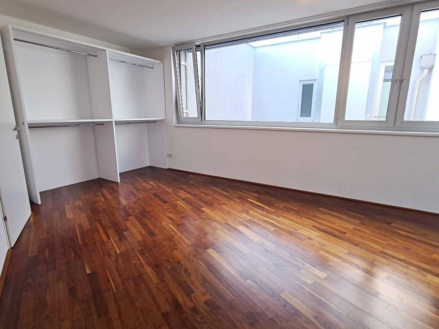Schlafzimmer mit Einbauschränken, großen Fenstern und dunklem Parkettboden.