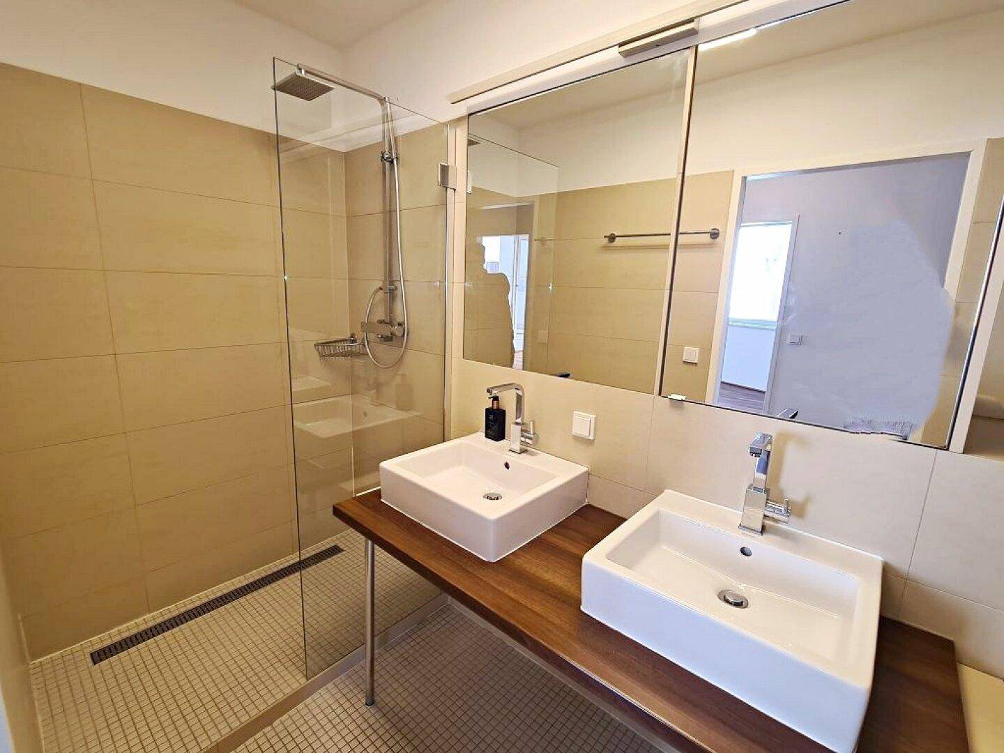 Modernes Badezimmer mit Doppelwaschbecken, großer Dusche und beigefarbenen Fliesen.